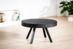 De Salontafel rond 74 cm, Lava, B341 vintage zwart, een ronde zwarte salontafel met vier schuine poten, staat op een lichte houten vloer in een moderne kamer met een witte muur, boeken op een plank en een groene potplant.