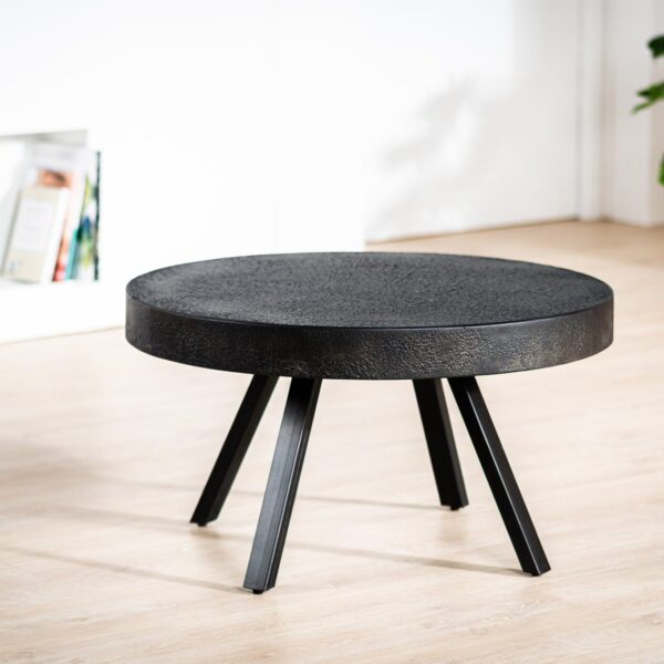 De Salontafel rond 74 cm, Lava, B341 vintage zwart, een ronde zwarte salontafel met vier schuine poten, staat op een lichte houten vloer in een moderne kamer met een witte muur, boeken op een plank en een groene potplant.