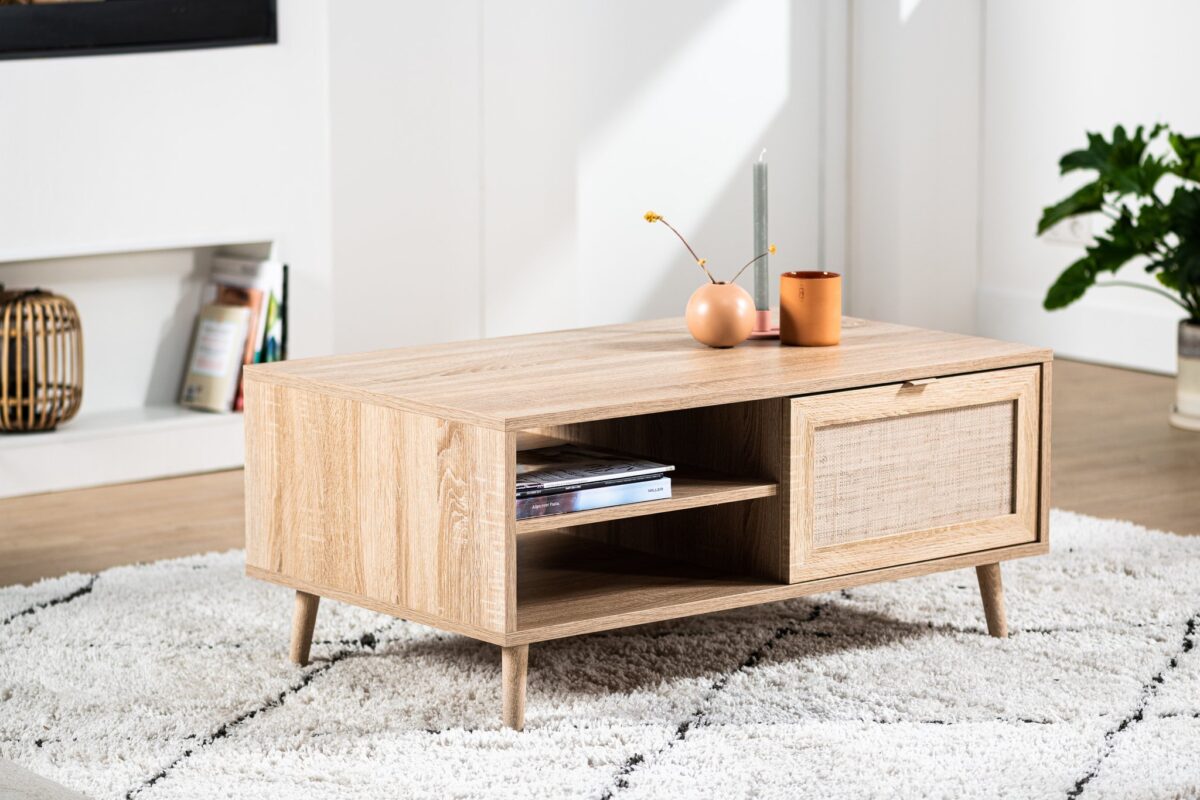 De Salontafel, 100x60 cm, M930 Sedona, heeft licht hout, schuine poten, een tijdschriftenplank en een lade met een getextureerd front. Een vaas, kaars en plant sieren het blad. Geplaatst op een wit vloerkleed met patroon.