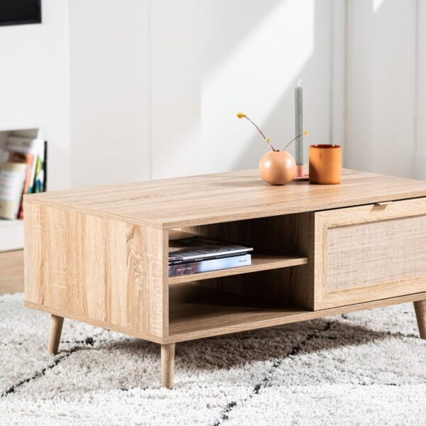 De Salontafel, 100x60 cm, M930 Sedona, heeft licht hout, schuine poten, een tijdschriftenplank en een lade met een getextureerd front. Een vaas, kaars en plant sieren het blad. Geplaatst op een wit vloerkleed met patroon.