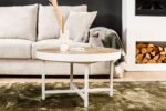 In een moderne woonkamer staat de Salontafel rond 74 cm, Lennox, L350 eiken salontafel, met daarop een vaas, kaars en boek. Daarachter staat een lichte bank met kussens en een deken. Een ingebouwde plank rechts is gevuld met brandhout.