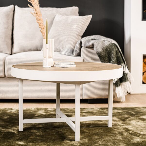 In een moderne woonkamer staat de Salontafel rond 74 cm, Lennox, L350 eiken salontafel, met daarop een vaas, kaars en boek. Daarachter staat een lichte bank met kussens en een deken. Een ingebouwde plank rechts is gevuld met brandhout.