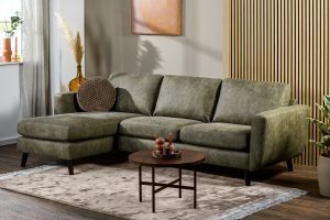 Een moderne woonkamer met een 3-zitsbank CL L+R, stof Savannah, S156 groene hoekbank, een ronde bruine salontafel, een beige vloerkleed, potplanten en een houten lattenwand. Kunst en natuurlijk licht dragen bij aan de gezellige en stijlvolle sfeer.