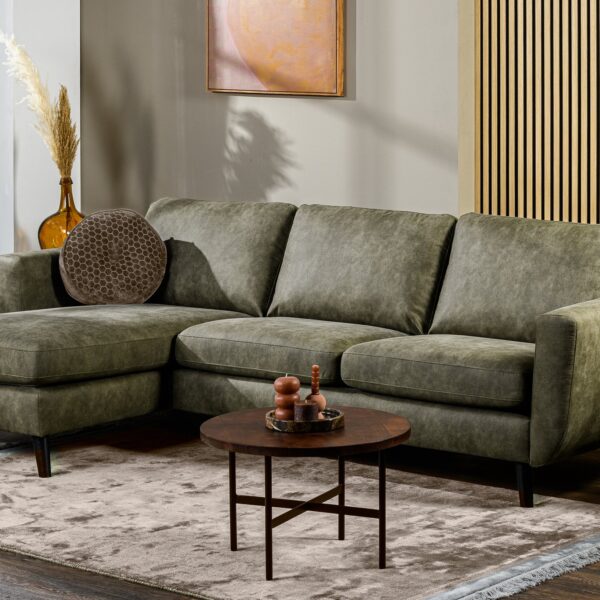 Een moderne woonkamer met een 3-zitsbank CL L+R, stof Savannah, S156 groene hoekbank, een ronde bruine salontafel, een beige vloerkleed, potplanten en een houten lattenwand. Kunst en natuurlijk licht dragen bij aan de gezellige en stijlvolle sfeer.