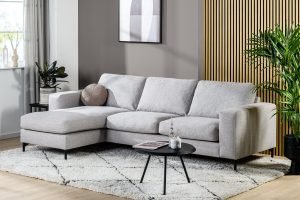 Moderne woonkamer met de 3-zitsbank CL L+R in Valente V311 grijs stof op een wit vloerkleed met patroon, een ronde zwarte salontafel, potplanten en wanddecoratie. Neutrale tinten en accentpanelen van hout.