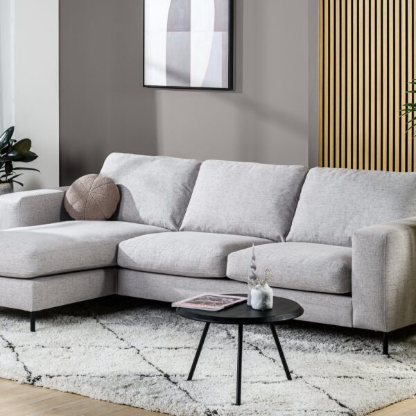Moderne woonkamer met de 3-zitsbank CL L+R in Valente V311 grijs stof op een wit vloerkleed met patroon, een ronde zwarte salontafel, potplanten en wanddecoratie. Neutrale tinten en accentpanelen van hout.