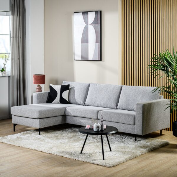 Een moderne woonkamer met de 3-zitsbank CL L+R in de stof Chloe C311 grijs op een wit vloerkleed, gecombineerd met een ronde, zwarte salontafel, een roze lamp, abstracte wandkunst en een hoge groene plant bij een accentmuur met houten lambrisering.