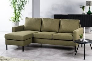 Een eigentijdse driezitsbank CL L+R in Dillon D156 groene stof met drie kussens en een chaise longue links, vormt de basis van een lichte woonkamer. Erachter staan een ronde zwarte bijzettafel met decoratie en planten, en een zwarte kast met vazen.
