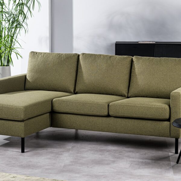 Een eigentijdse driezitsbank CL L+R in Dillon D156 groene stof met drie kussens en een chaise longue links, vormt de basis van een lichte woonkamer. Erachter staan een ronde zwarte bijzettafel met decoratie en planten, en een zwarte kast met vazen.
