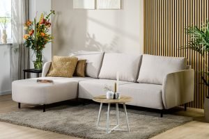 In een moderne woonkamer staat de 3-zitsbank CL L+R in de beige stof Urban U460, met daarbij twee sierkussens, een ronde salontafel met kaarsen en vazen, een grijs vloerkleed, decoratieve planten tegen een houten wand en bloemen bij het raam.