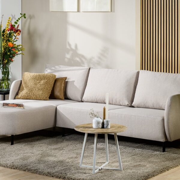 In een moderne woonkamer staat de 3-zitsbank CL L+R in de beige stof Urban U460, met daarbij twee sierkussens, een ronde salontafel met kaarsen en vazen, een grijs vloerkleed, decoratieve planten tegen een houten wand en bloemen bij het raam.