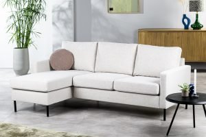 Moderne woonkamer met de 3-zitsbank CL L+R in Valente V920 naturel, een rond bruin kussen, een zwarte ronde salontafel, een houten dressoir, een grote potplant en decoratie tegen een lichtgrijze muur.