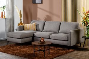 Moderne woonkamer met de 3-zitsbank CL L+R in de kleur Woven W520 taupe, met gestreepte kussens, een ronde houten salontafel op een bruin tapijt, decoratieve planten, houten lambrisering en bruine muren, en zonlicht door transparante gordijnen.
