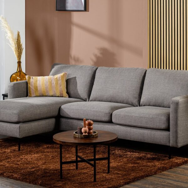 Moderne woonkamer met de 3-zitsbank CL L+R in de kleur Woven W520 taupe, met gestreepte kussens, een ronde houten salontafel op een bruin tapijt, decoratieve planten, houten lambrisering en bruine muren, en zonlicht door transparante gordijnen.