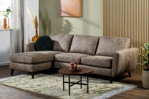 Een moderne woonkamer met de 3-zitsbank CL L+R in Savannah S520 taupe, zwarte poten, een ronde houten salontafel, een lichtgroen tapijt, planten, decoratieve vazen, wandkunst en zonlicht dat door het raam naar binnen stroomt.