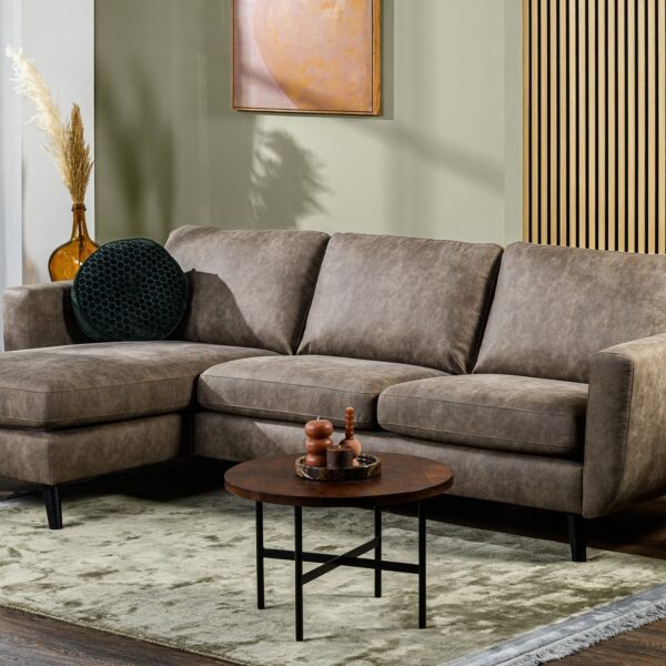 Een moderne woonkamer met de 3-zitsbank CL L+R in Savannah S520 taupe, zwarte poten, een ronde houten salontafel, een lichtgroen tapijt, planten, decoratieve vazen, wandkunst en zonlicht dat door het raam naar binnen stroomt.