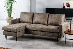 De 3-zitsbank CL L+R, stof Savannah, S520 taupe - een moderne taupekleurige hoekbank met drie zitplaatsen en een verlengde loungehoek - staat in een lichte woonkamer met een lichtgrijze vloer, een potplant en een ronde houten salontafel.