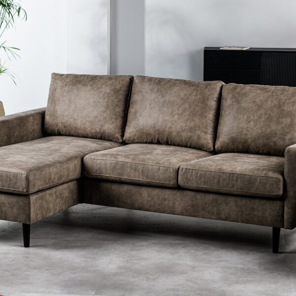 De 3-zitsbank CL L+R, stof Savannah, S520 taupe - een moderne taupekleurige hoekbank met drie zitplaatsen en een verlengde loungehoek - staat in een lichte woonkamer met een lichtgrijze vloer, een potplant en een ronde houten salontafel.