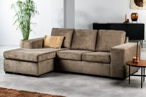 De 3-zitsbank CL L+R in de stof Hotel Chique H430 bruin heeft een modern modulair ontwerp met een chaise longue en een geel gestreept kussen. De bank staat in een lichte woonkamer met een plant, een ronde houten tafel en een dressoir.