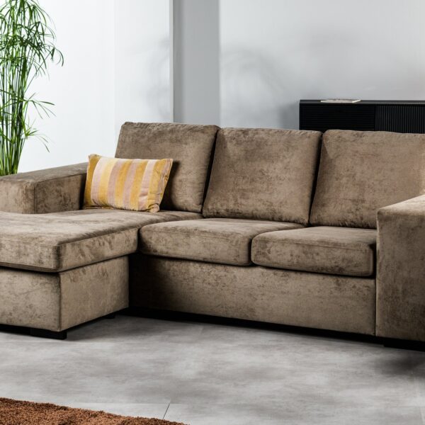 De 3-zitsbank CL L+R in de stof Hotel Chique H430 bruin heeft een modern modulair ontwerp met een chaise longue en een geel gestreept kussen. De bank staat in een lichte woonkamer met een plant, een ronde houten tafel en een dressoir.