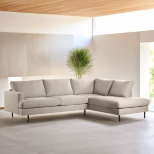 Een moderne woonkamer met de 3-zits hoek rechts, stof Bjorn 24, W460 beige hoekbank met slanke donkere poten en grote kussens. De ruimte is minimalistisch ingericht, heeft een potplant en veel natuurlijk licht dankzij de grote ramen.