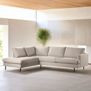 In de moderne woonkamer staat de beige L-vormige hoekbank met 3 zits hoek links, stof Bjorn 24, W460 op een lichte vloer, met een grote groene plant en natuurlijk licht dat door de hoge ramen op de achtergrond naar binnen stroomt.