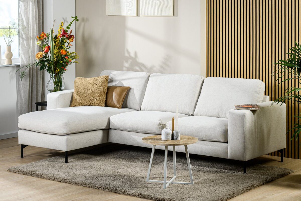 Moderne woonkamer met de 3-zitsbank CL L+R in de stof Valente V920 naturel, twee decoratieve kussens, een kleine ronde salontafel met kaarsen, een vaas met kleurrijke bloemen, een grijs vloerkleed en houten lambrisering.