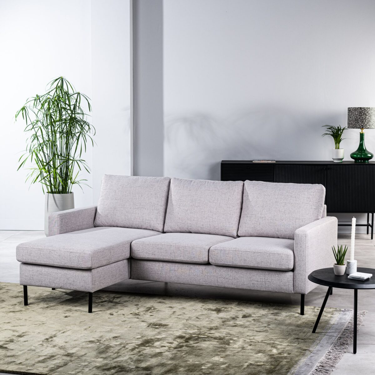 Moderne woonkamer met de 3-zitsbank CL L+R, stof Valente, V311 grijze hoekbank, groen vloerkleed, ronde zwarte bijzettafel met kleine plantjes, hoge potplant en zwart dressoir met decoratie tegen een witte muur.