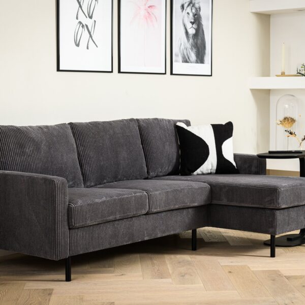 Moderne woonkamer met de 3-zitsbank CL L+R in RIB230 antraciet ribstof, een zwart-wit accentkussen, een lichte visgraatvloer, planten, drie ingelijste kunstwerken op een crèmekleurige muur en minimalistische decoratie op witte planken.