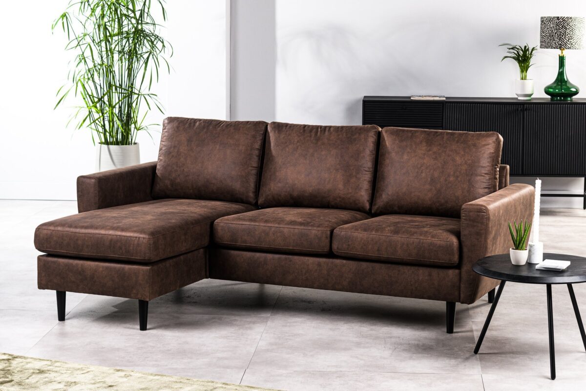 Een moderne woonkamer met een 3-zitsbank CL L+R in Savannah S430 bruin, zwarte poten, een klein rond zwart bijzettafeltje met een potplant en afstandsbediening, en decoratieve planten op de achtergrond.
