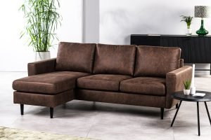 Een moderne woonkamer met een 3-zitsbank CL L+R in Savannah S430 bruin, zwarte poten, een klein rond zwart bijzettafeltje met een potplant en afstandsbediening, en decoratieve planten op de achtergrond.