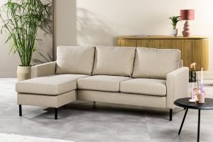 Een 3-zitsbank CL L+R in de beige stof Dillon D460 staat in een lichte woonkamer, gecombineerd met een potplant, een ronde zwarte salontafel en een dressoir met decoratieve items.