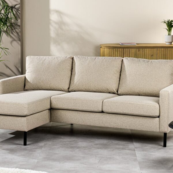 Een 3-zitsbank CL L+R in de beige stof Dillon D460 staat in een lichte woonkamer, gecombineerd met een potplant, een ronde zwarte salontafel en een dressoir met decoratieve items.