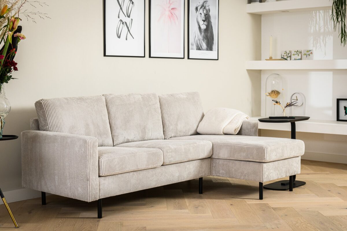 Een 3-zitsbank CL L+R, stof RIB, RIB920 naturel staat in een moderne woonkamer met een ingelijste muurschildering, witte planken en een klein zwart bijzettafeltje met een kussen.