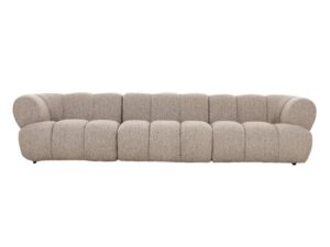 Een lange, moderne beige bank met een zachte, gestructureerde stof. De bank heeft zes grote, ronde kussens die de rugleuning en zitting vormen, wat een gezellige en uitnodigende uitstraling creëert. Hij staat laag bij de grond.