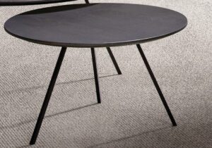 Een strak, rond, zwart metalen bijzettafeltje met drie slanke poten staat op een beige tapijt.