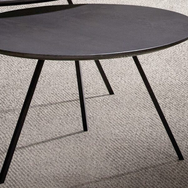 De Izola SALONTAFEL120 E02M, een ronde, zwarte bijzettafel met drie poten, staat op een licht gestructureerd tapijt. Op de achtergrond is nog een soortgelijke tafel gedeeltelijk zichtbaar.