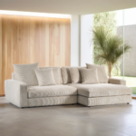 Een 3-zits bank CL L+R in Lincoln 83 stof, kleur RIB920 naturel, staat in een moderne woonkamer met grote ramen, een houten accentmuur en een palmplant. Natuurlijk licht vult de minimalistische ruimte.