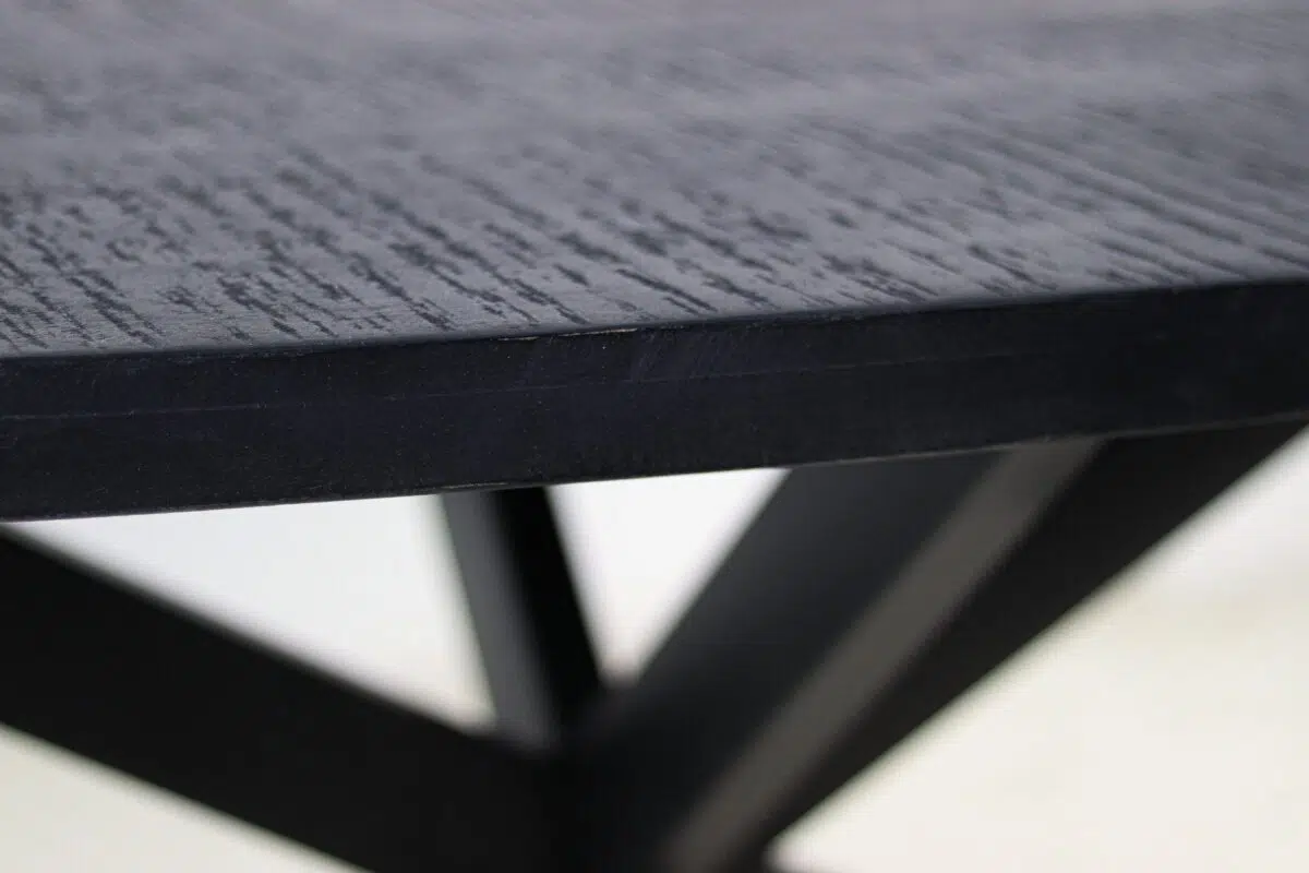 Close-up van een zwarte houten tafelrand met een gestructureerd oppervlak, ondersteund door hoekige metalen poten. De achtergrond is wazig, waardoor het ontwerp en vakmanschap van de tafel worden benadrukt.