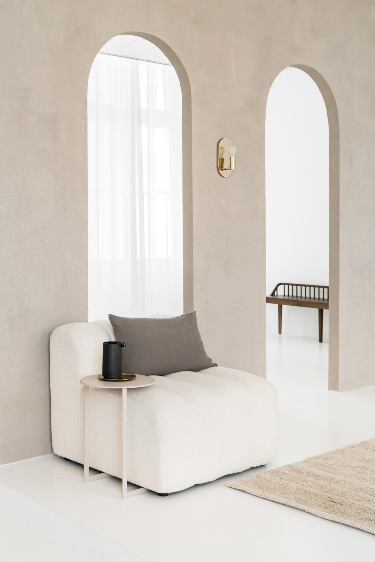 Een minimalistische leefruimte met een crèmekleurige stoel, grijs kussen en een klein rond bijzettafeltje met een zwarte vaas. Twee gebogen deuropeningen en een houten bankje op de achtergrond, met een neutraal kleed op de vloer.