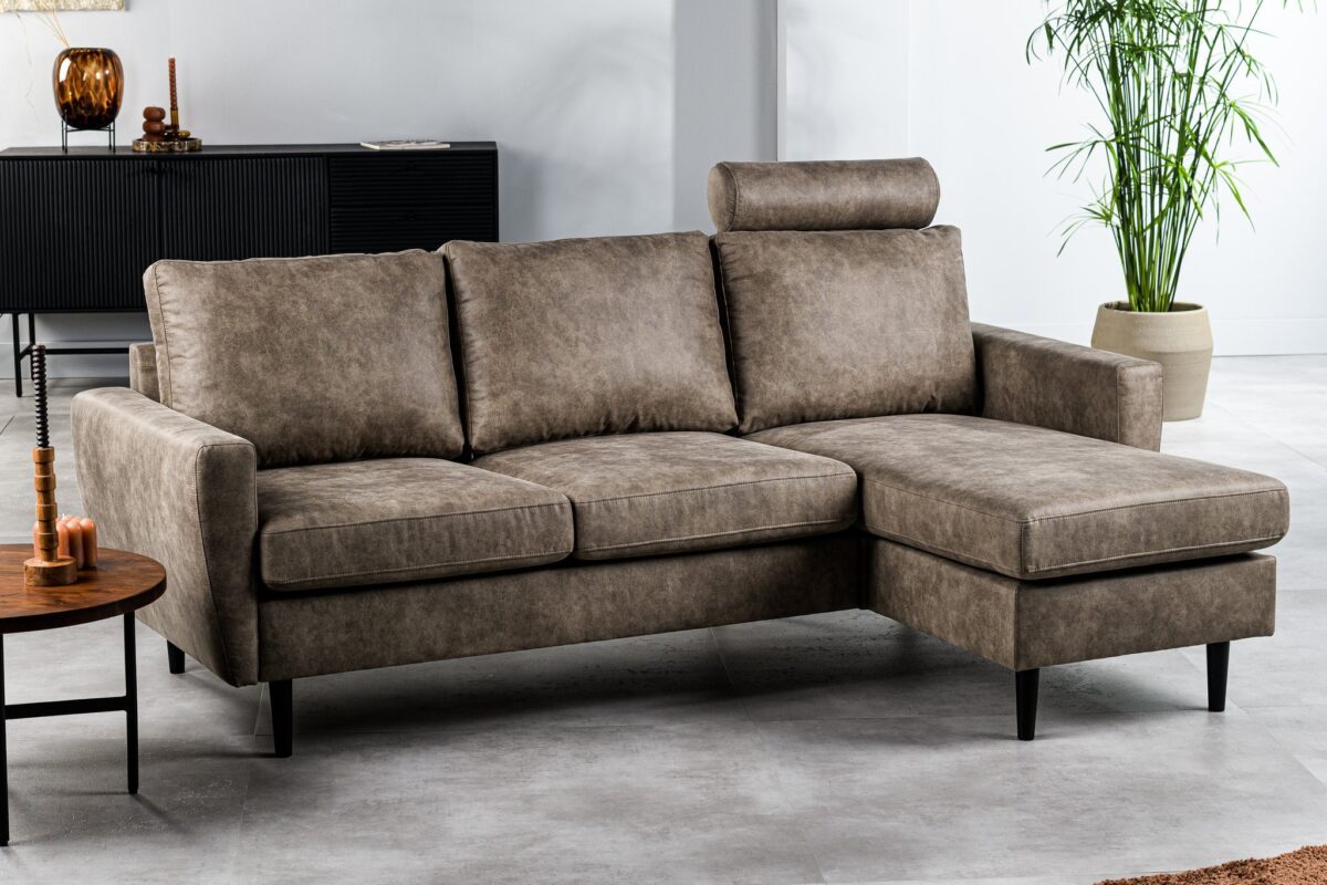 Een moderne woonkamer heeft een bruine L-vormige bank met een hoofdsteun, geplaatst op een lichtgrijze vloer. Er is een zwart dressoir met decoratieve items op de achtergrond en een groene potplant aan de rechterkant.