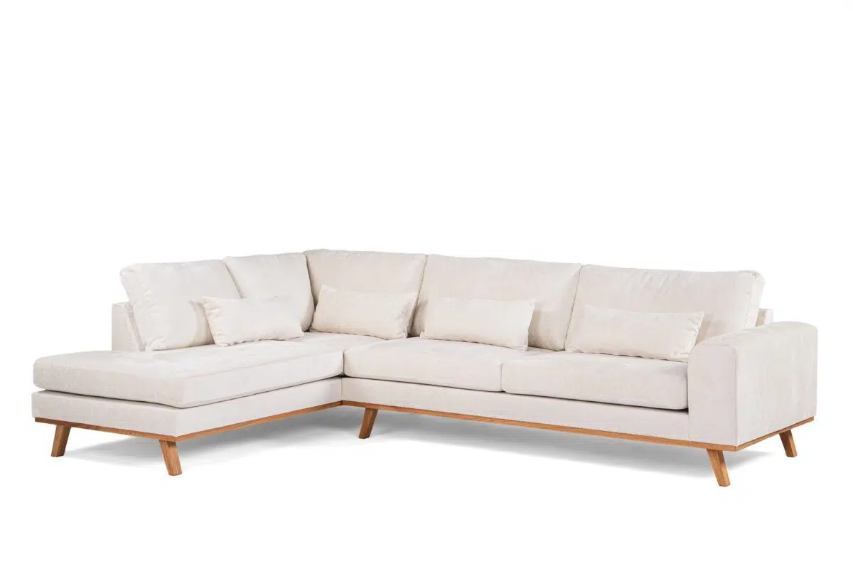 Een lichtbeige L-vormige sectionele bank met houten poten op een witte achtergrond. De bank heeft drie zitkussens, een chaise longue aan de linkerkant en vijf bijpassende sierkussens.