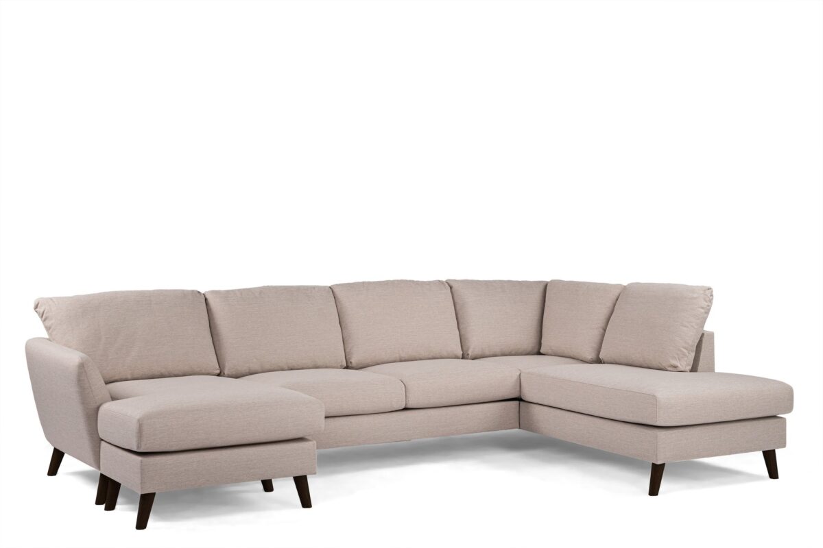 Een moderne, beige sectionele bank met een chaise longue aan de linkerkant en een schuine verlenging aan de rechterkant. Het heeft gewatteerde zittingen en een lage rugleuning, ondersteund door donkere houten poten. Het ontwerp is minimalistisch en eigentijds.