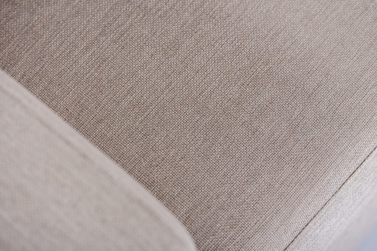 Close-up van een beige stoffen bank die de getextureerde bekleding met fijne, gelijkmatige stiksels laat zien. De afbeelding benadrukt het strakke, geweven patroon en de zachte uitstraling van de stof.