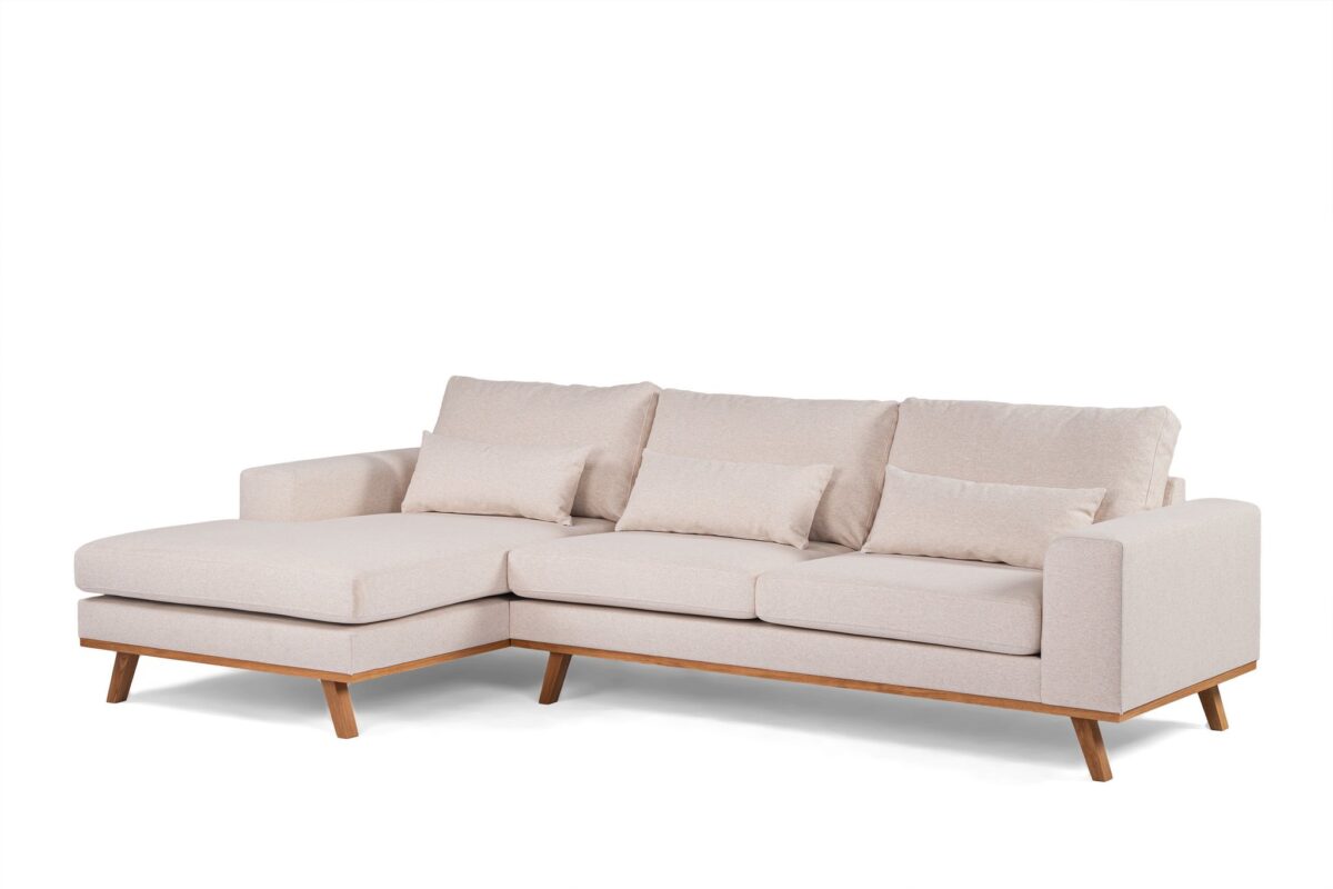 Een beige L-vormige sectionele bank met houten poten. Het heeft drie grote zitkussens, drie rugkussens en twee kleinere sierkussens. De bank heeft een minimalistisch, modern design met strakke lijnen.