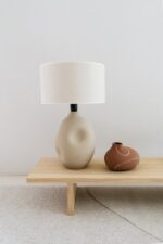 Een minimalistische scène met een beige keramische lamp met een abstracte vorm en witte kap op een houten bank naast een kleine bruine vaas met witte lineaire accenten. De achtergrond is een neutrale muur en vloer.
