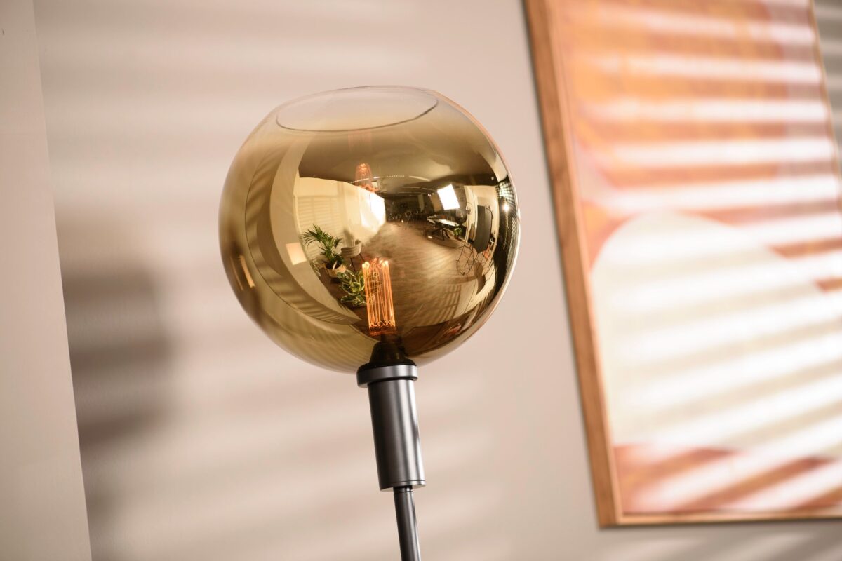 Een moderne staande lamp met een bolvormige, amberkleurige glazen kap en een zichtbare gloeilamp staat tegen een beige muur. Een ingelijst kunstwerk met horizontale lijnen is gedeeltelijk zichtbaar, wat warm, natuurlijk licht reflecteert.