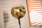 Een moderne staande lamp met een bolvormige, amberkleurige glazen kap en een zichtbare gloeilamp staat tegen een beige muur. Een ingelijst kunstwerk met horizontale lijnen is gedeeltelijk zichtbaar, wat warm, natuurlijk licht reflecteert.