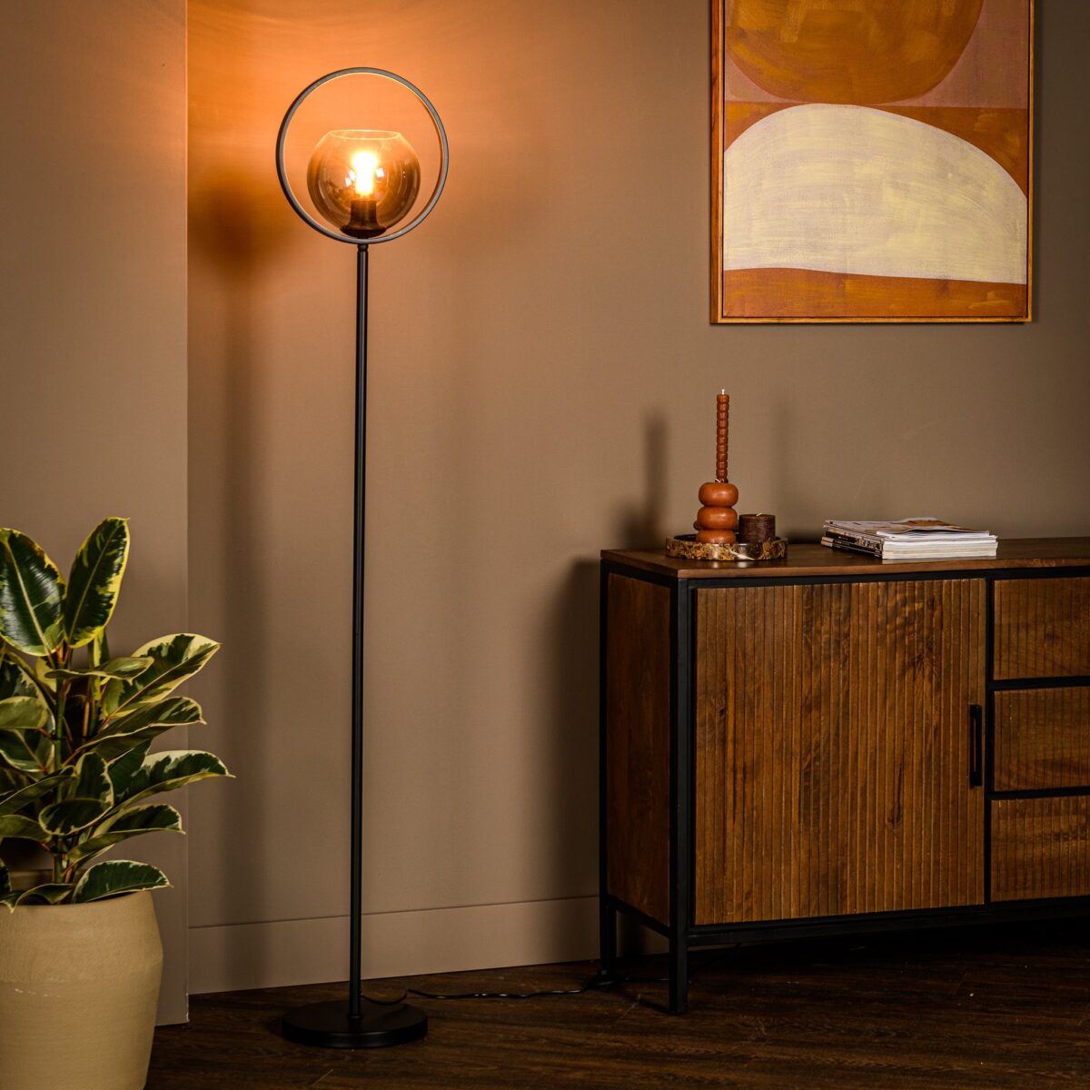 Een moderne woonkamer heeft een hoge staande lamp met een glazen bolvormige kap. Een houten kast met boeken en decoratieve items staat tegen een taupe muur versierd met abstracte kunst. Een potplant staat op de houten vloer.