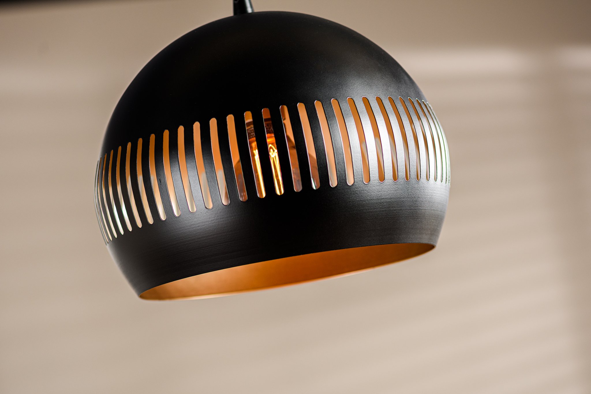 Een close-up van een moderne, zwarte hanglamp met verticale uitsparingen die het midden omcirkelen, waardoor een warme gloed van de lamp erin zichtbaar wordt. De achtergrond is een zacht wazige neutrale setting, wat een binnenomgeving suggereert.